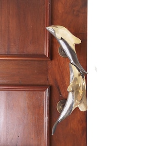 Puede incluir: Un par de tiradores de puerta de delfín de latón, uno apilado sobre el otro, montados en una puerta de madera.
