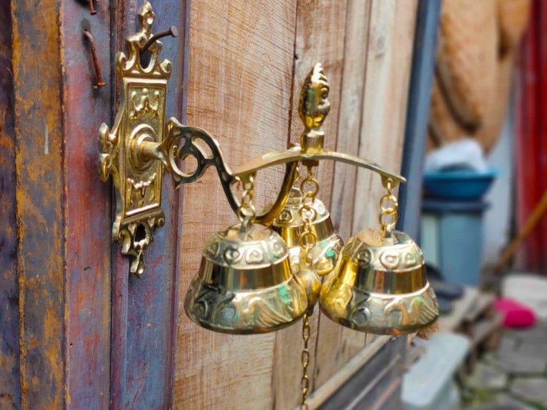 May include: A gold-toned door chime with three bells hanging from a decorative metal bracket. The bells are ornate and have intricate designs.