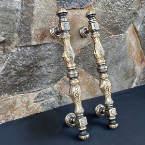 May include: Two antique-style door handles with an ornate design. The handles are made of a gold-colored metal with dark accents. The handles have a long, curved shape with decorative floral and scrollwork details. The handles are shown against a textured stone background.