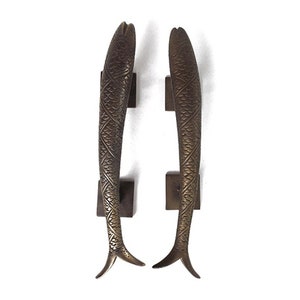 May include: A pair of bronze-toned door handles, each sculpted as a fish with detailed scales and fins. They are mounted on dark brown, square plates. The fish handles are mirror images of each other.