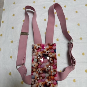 Peut inclure: Bretelles roses avec un porte-cartes rectangulaire orné d'un motif de chiots et de cœurs. Le porte-cartes a une fenêtre en plastique transparent. Les bretelles ont des fermoirs métalliques et des sangles réglables.