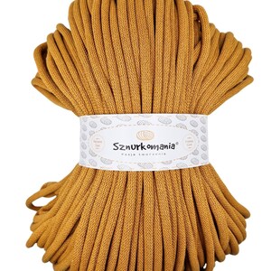 May include: A bundle of mustard-coloured macrame cord, tightly wound and secured with a white label. The label reads "Sznurkomania" and "Pasja tworzenia". The cord is a thick, textured material, suitable for various crafting projects.