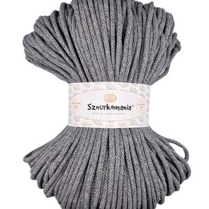 Può includere: Un cordoncino di cotone grigio, avvolto a forma di palla, con un'etichetta bianca su cui è scritto "Sznurkomania Pasja tworzenia" e un'immagine piccola di un gomitolo di lana.