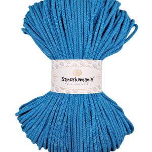 May include: A vibrant blue skein of macrame cord. The cord is tightly wound and held together by a white label with the text "Sznurkomania" and "Pasja tworzenia". The cord appears to be made of a soft, durable material, ideal for crafting projects.