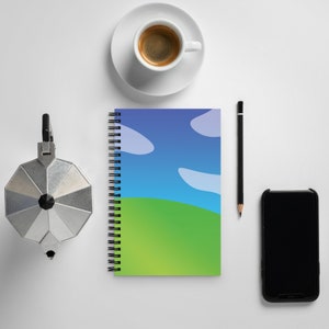 May include: A spiral bound notebook with a blue, green, and white landscape design. The notebook is on a white surface with a coffee cup, a pencil, and a smartphone.