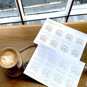 Op de afbeelding: Een latte in een heldere glazen beker met latte art, naast twee vellen papier met lijntekeningen van een torso en de tekst "Pineapple Studio x rika_akir ink Tasting". De items liggen op een houten oppervlak.