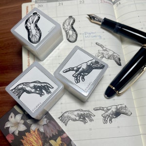 May include: Four white square rubber stamps with black ink. The stamps feature different images, including a hand reaching out, a hand with a finger pointing, and a stylized image of a clam. The stamps are on a white page with a calendar grid and the text "Digital Marketing".