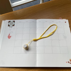May include: Open calendar with the month of October displayed. A small, round, cream-colored object with a yellow strap lies on the calendar. The calendar is open on a wooden table near a window.