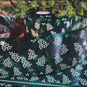 May include: A roll of green washi tape with a white floral pattern. The flowers are made up of small white circles and the stems are a dark green. The tape has a subtle shimmer.