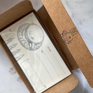 Puede incluir: Un juego de papelería en una caja de cartón marrón. La papelería presenta un diseño de pluma y tinta. La caja tiene una ilustración de un conejito en el lateral. La papelería está empaquetada en una funda de plástico transparente.