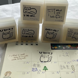 Puede incluir: Una colección de pequeños sellos cuadrados de color blanquecino con ilustraciones de gatos negros y frases motivadoras como "U ROCK!" y "AMAZING!". Un calendario con notas manuscritas y un dibujo de gato.