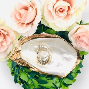 May include: A gold-rimmed oyster shell ring dish with a white interior, surrounded by pink roses and green foliage. A gold ring is resting in the dish.