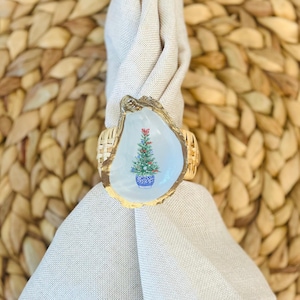 May include: A white oyster shell napkin ring with a gold rim and a painted Christmas tree design. The tree is green with red and blue ornaments and is in a blue and white pot. The napkin ring is on a white linen napkin.