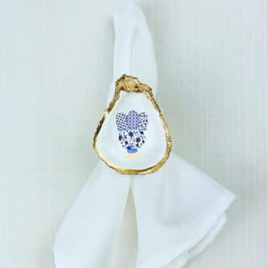 May include: Gold-rimmed oyster shell napkin ring with a blue and white floral design. The design features a potted plant with blue flowers and white leaves.