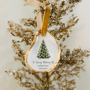 May include: A white oyster shell ornament with a gold trim, featuring a green Christmas tree with gold crosses. The ornament reads "In Loving Memory Of Johnathon 1934-2000".