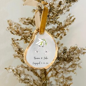 May include: White oyster shell ornament with gold trim, featuring two martini glasses with olives and the text "Briana & Jake Engaged 7.13.24".