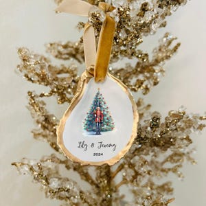 May include: A white oyster shell Christmas ornament with gold trim, featuring a hand-painted illustration of a Christmas tree with a nutcracker standing in front of it. The ornament is personalized with the text "Lily & Jeremy 2024".