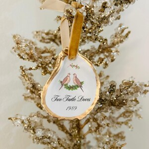 May include: An oyster shell Christmas ornament with a gold rim and a cream-colored interior. The ornament features an illustration of two turtle doves and the text "Two Turtle Doves 1989". A gold ribbon is attached for hanging.