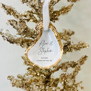 May include: White oyster shell ornament with gold trim. The ornament is decorated with the names "Alex & Stephen" and the date "5/11/24 CHARLESTON, SC".
