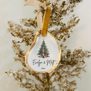 May include: A white oyster shell Christmas ornament with gold trim. The ornament features a watercolor illustration of a Christmas tree with a nutcracker standing in front of it. The text "Evelyn & Matt 2024" is written below the tree.