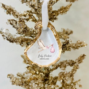 May include: A white oyster shell ornament with gold trim, featuring a watercolor illustration of a stork carrying a pink baby bundle. The text "Baby Pearlman Arriving 2025" is printed on the shell.