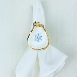 May include: A white oyster shell napkin ring with a gold-painted edge and a blue snowflake design in the center. The napkin ring is holding a white linen napkin. The design is perfect for winter or holiday table settings.