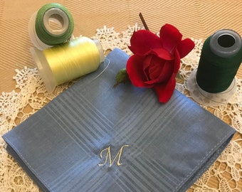 May include: A light blue handkerchief with a white embroidered letter "M" in the center. The handkerchief is on a white lace doily with spools of green and yellow thread and a red rose.
