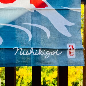 Pu&ograve; includere: Uno stendardo di stoffa blu con un disegno di pesce koi bianco e il testo "Nishikigoi" in corsivo bianco. Un timbro rosso con il testo "Atsu" si trova nell'angolo in basso a destra.