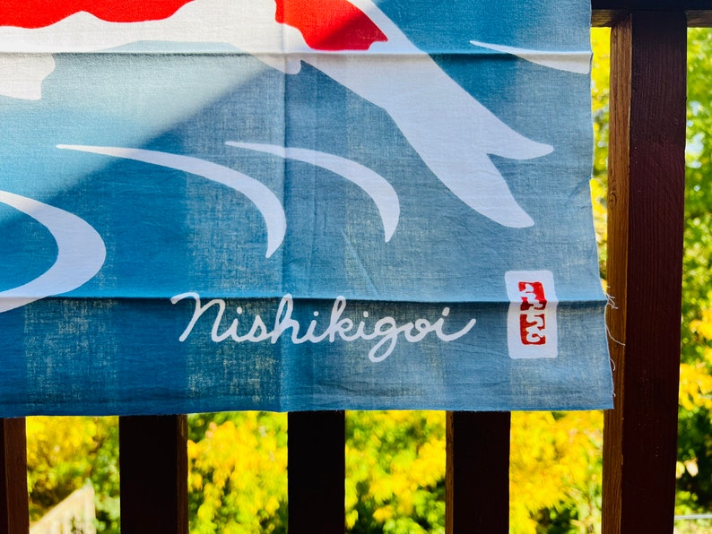 Pu&ograve; includere: Uno stendardo di stoffa blu con un disegno di pesce koi bianco e il testo "Nishikigoi" in corsivo bianco. Un timbro rosso con il testo "Atsu" si trova nell'angolo in basso a destra.