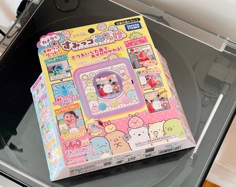 May include: A pink and purple toy camera with a screen that shows a cartoon character. The camera is surrounded by other cartoon characters. The box is labeled "Sumikko Gurashi" and "Takara Tomy".