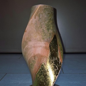 May include: A tall, abstract ceramic vase with a unique, twisted shape. The vase features a blend of colors, including green, peach, and gold leaf accents. It stands on a small, round base, showcasing an artistic and decorative design.