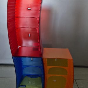 Könnte beinhalten: Ein modulares Aufbewahrungssystem für Compact Discs aus durchscheinendem Kunststoff in Rot, Blau und Orange. Die Struktur besteht aus drei geschwungenen, stapelbaren Elementen mit internen Schlitzen zur Medienorganisation. Der blaue Teil trägt das Etikett koziol.