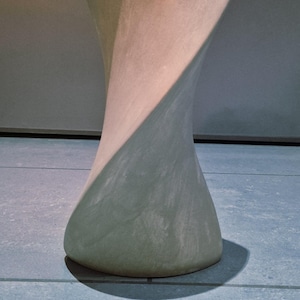 May include: A light gray, sculptural, hourglass-shaped object with a textured surface. The object is set against a neutral background and sits on a blue tiled floor. The design features a twist.