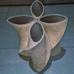 May include: A unique, abstract sculpture with four interconnected, petal-like forms. The sculpture is a light beige color with a gray rim around the openings. The design is modern and artistic, suitable for home decor.