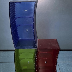 Può includere: Un set di quattro unità di stoccaggio in plastica colorata e curva, blu, verde e rossa. Ogni unità ha una parte superiore strutturata e una piccola apertura. Le unità sono impilate, creando una forma unica.