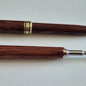 May include: Two wooden pens with gold-colored accents. One pen is a ballpoint pen with a gold tip, and the other is a stylus pen. The pens are a dark brown color and have the word "Zetaminch" engraved on the side.