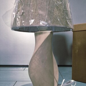 May include: A beige table lamp with a textured lampshade covered in plastic. The lamp has a curved, vase-like base. A cardboard box is visible in the background. The lamp is on a tiled floor.