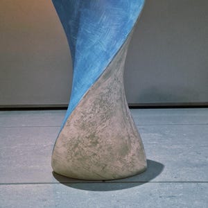 May include: A decorative, hourglass-shaped vase. The top half is painted blue, while the bottom half is a textured, off-white color. The vase is set on a blue tiled surface, with a shadow cast to the right.