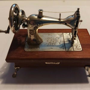 Pode incluir: Uma máquina de costura prateada em miniatura com manivela e base de madeira castanha escura. A máquina de costura tem detalhes intrincados e assenta numa base retangular com detalhes metálicos decorativos. A base tem pequenos pés ornamentados.