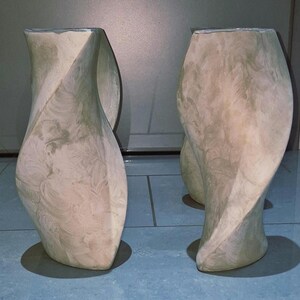 May include: Two beige, abstract, sculptural vases with a twisted design. The vases have a textured surface and a wide base. The overall shape is elongated and organic, with a neutral color palette.