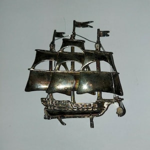 Puede incluir: Broche plateado con forma de barco con tres mástiles y velas ondeantes. El broche presenta detalles intrincados de la estructura del barco, incluyendo el casco y el aparejo. Las banderas en la parte superior de cada mástil son visibles. El broche es probablemente de metal.