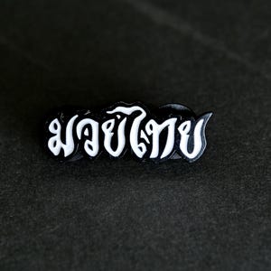 May include: A black pin with the Thai text "มวยไทย" in white, stylized lettering. The pin is rectangular with rounded edges and is set against a dark, textured background.