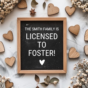 May include: A wooden chalkboard with the text "The Smith Family is Licensed to Foster!" surrounded by small wooden hearts and white flowers.