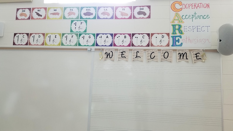 Pu&ograve; includere: Una lavagna bianca con uno striscione di benvenuto e un cartello che dice "CARE" con le parole "Cooperazione", "Accettazione", "Rispetto" e "Entusiasmo" scritte sotto. Ci sono anche note musicali e segni delle mani sul muro.