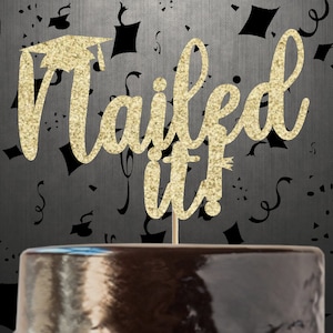 May include: A gold glitter cake topper with the words "Nailed it!" and a graduation cap. The topper is on a wooden stick, placed on top of a dark cake. The background is dark gray with black confetti.