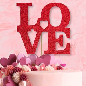 Pode incluir: Topper de bolo vermelho com glitter que diz "LOVE" com um pequeno coração no meio do "O".