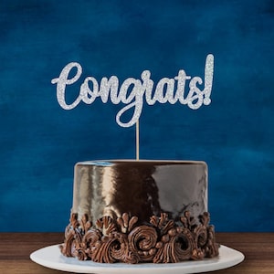 May include: A chocolate cake with a silver glitter "Congrats!" cake topper. The cake is decorated with chocolate frosting and swirls.