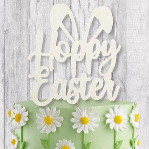 May include: A cake topper with the words "Hoppy Easter" in a cursive font, with bunny ears above the text. The cake is decorated with white daisies and green frosting. The background is a light gray wooden surface.