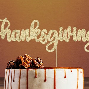 Peut inclure: Un topper de gâteau en paillettes dorées avec le mot "Thanksgiving" en écriture cursive. Le topper est placé sur un gâteau blanc sur lequel est versée une sauce au caramel et surmonté de noix enrobées de caramel.