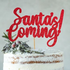 Może przedstawiać: Biały tort z czerwonym, błyszczącym topperem, na którym widnieje napis "Santa's Coming!" Tort jest ozdobiony gałązkami rozmarynu i czerwonymi jagodami.